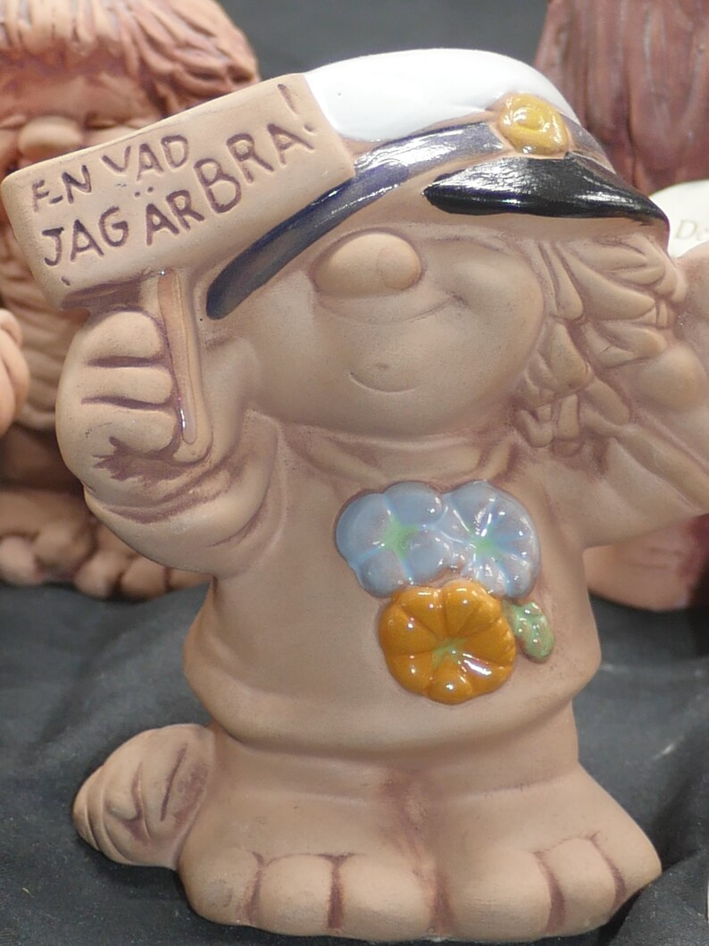 Pu&ograve; includere: Una statuetta in ceramica di un gnomo sorridente che indossa un cappello da marinaio blu e bianco. Lo gnomo tiene un cartello con su scritto "EN VAD JAG &Auml;R BRA!" Lo gnomo indossa una camicia marrone con fiori blu e arancioni.