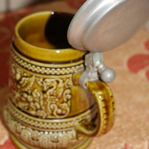 Może przedstawiać: Brązowy ceramiczny kufel do piwa z metalową pokrywką i szczeg&oacute;łowym wzorem kwiatowym. Kufel ma uchwyt i okrągłą podstawę.