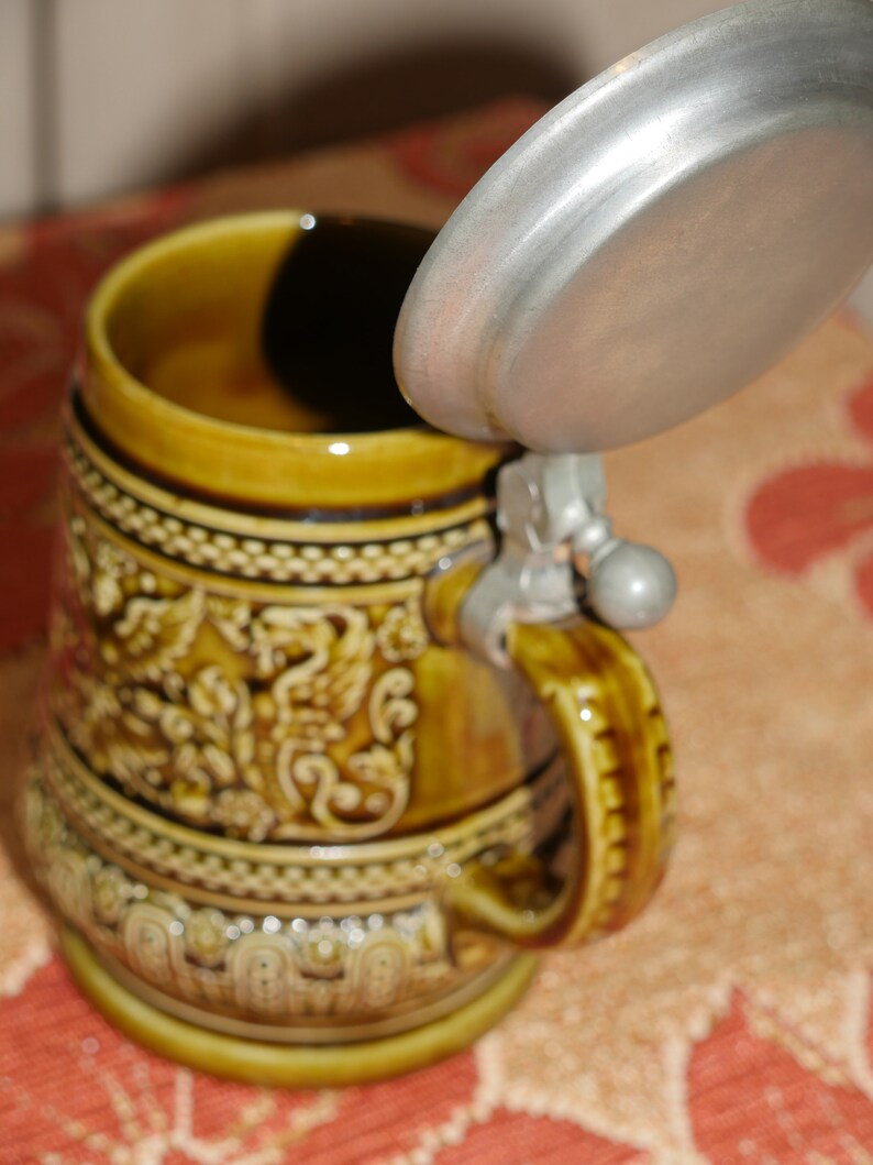 Może przedstawiać: Brązowy ceramiczny kufel do piwa z metalową pokrywką i szczeg&oacute;łowym wzorem kwiatowym. Kufel ma uchwyt i okrągłą podstawę.