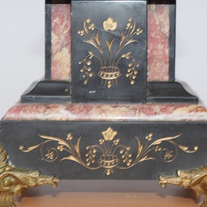 May include: Black marble clock with gold accents and floral designs. The clock has a square base with a pink marble inlay and sits on gold-toned feet.