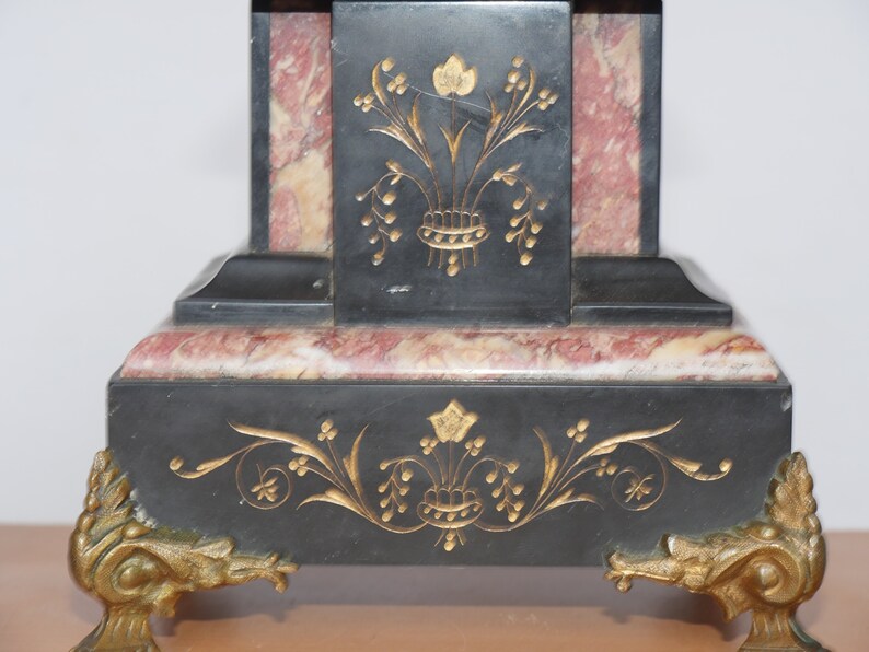 May include: Black marble clock with gold accents and floral designs. The clock has a square base with a pink marble inlay and sits on gold-toned feet.