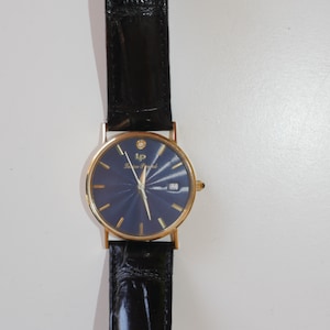 May include: A gold-tone wristwatch with a blue face and a black leather band. The watch has a diamond at the 12 o'clock position and the words "Lauren  Frank" on the face.