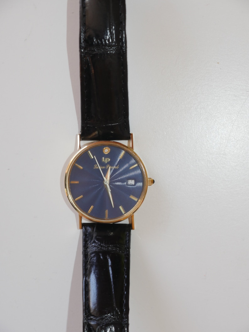 May include: A gold-tone wristwatch with a blue face and a black leather band. The watch has a diamond at the 12 o'clock position and the words "Lauren  Frank" on the face.