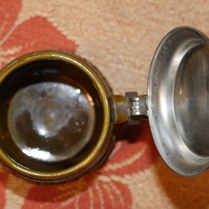 Może przedstawiać: Brązowy ceramiczny kufel do piwa z pokrywką z cyny. Kufel ma szczeg&oacute;łowy wz&oacute;r, a pokrywka ma zawiasowy uchwyt.