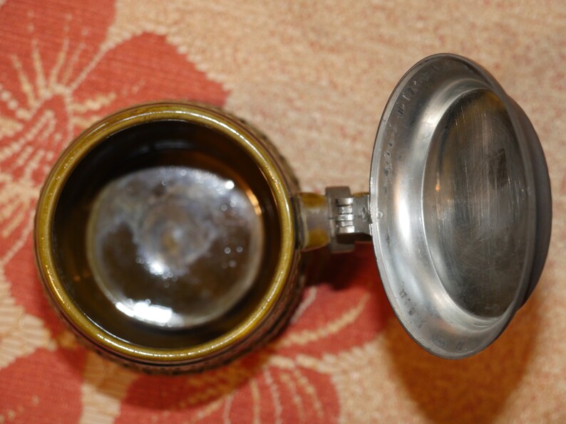 Może przedstawiać: Brązowy ceramiczny kufel do piwa z pokrywką z cyny. Kufel ma szczeg&oacute;łowy wz&oacute;r, a pokrywka ma zawiasowy uchwyt.