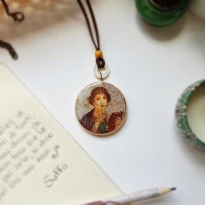 May include: A round pendant necklace with a brown cord. The pendant features a portrait of a person holding a book. A pencil and a notepad with handwritten text are in the background.