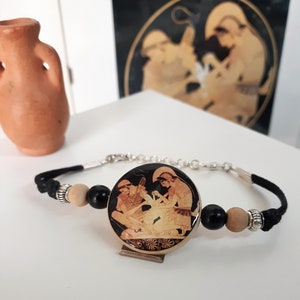 Puede incluir: Un brazalete de cord&oacute;n negro con un dije redondo de madera con una imagen en blanco y negro de dos figuras de estilo griego.
