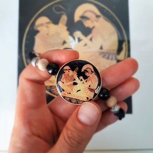 Puede incluir: Un brazalete negro y marr&oacute;n con un colgante redondo que presenta una imagen en blanco y negro de dos figuras en una escena de la mitolog&iacute;a griega. El colgante est&aacute; rodeado de cuentas de madera.