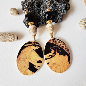 May include: A pair of dangle earrings with black and gold beads and oval-shaped pendants featuring black and white images of Greek figures. The pendants are made of wood and have a white background with a black border.