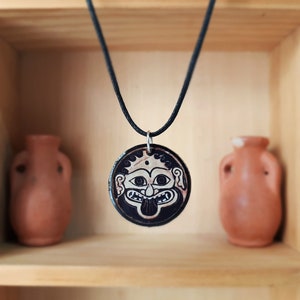 May include: A black cord necklace with a round ceramic pendant featuring a black and white illustration of a Gorgon's face. The pendant is set against a wooden background with two terracotta pots.