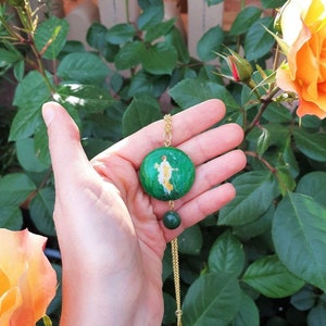 May include: A gold chain necklace with a green round pendant featuring a painted image of a person. The pendant is attached to a smaller green bead.