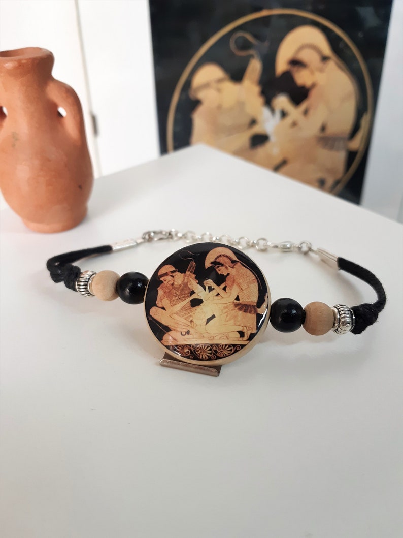 Puede incluir: Un brazalete de cord&oacute;n negro con un dije redondo de madera con una imagen en blanco y negro de dos figuras de estilo griego.