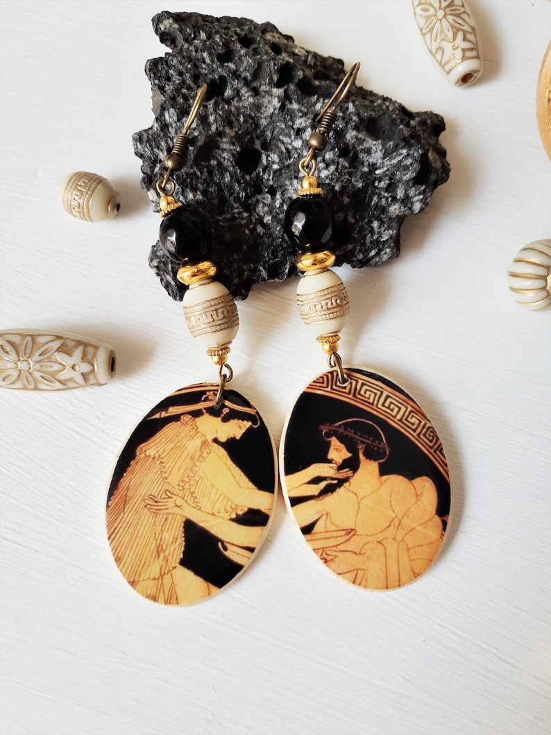 May include: A pair of dangle earrings with black and gold beads and oval-shaped pendants featuring black and white images of Greek figures. The pendants are made of wood and have a white background with a black border.