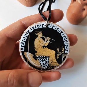 Peut inclure: Un collier pendentif rond avec un fond noir repr&eacute;sentant une sc&egrave;ne grecque classique d'une femme jouant de la fl&ucirc;te. Le pendentif a une bordure blanche avec un motif de cl&eacute; grecque et est sur un cordon noir.