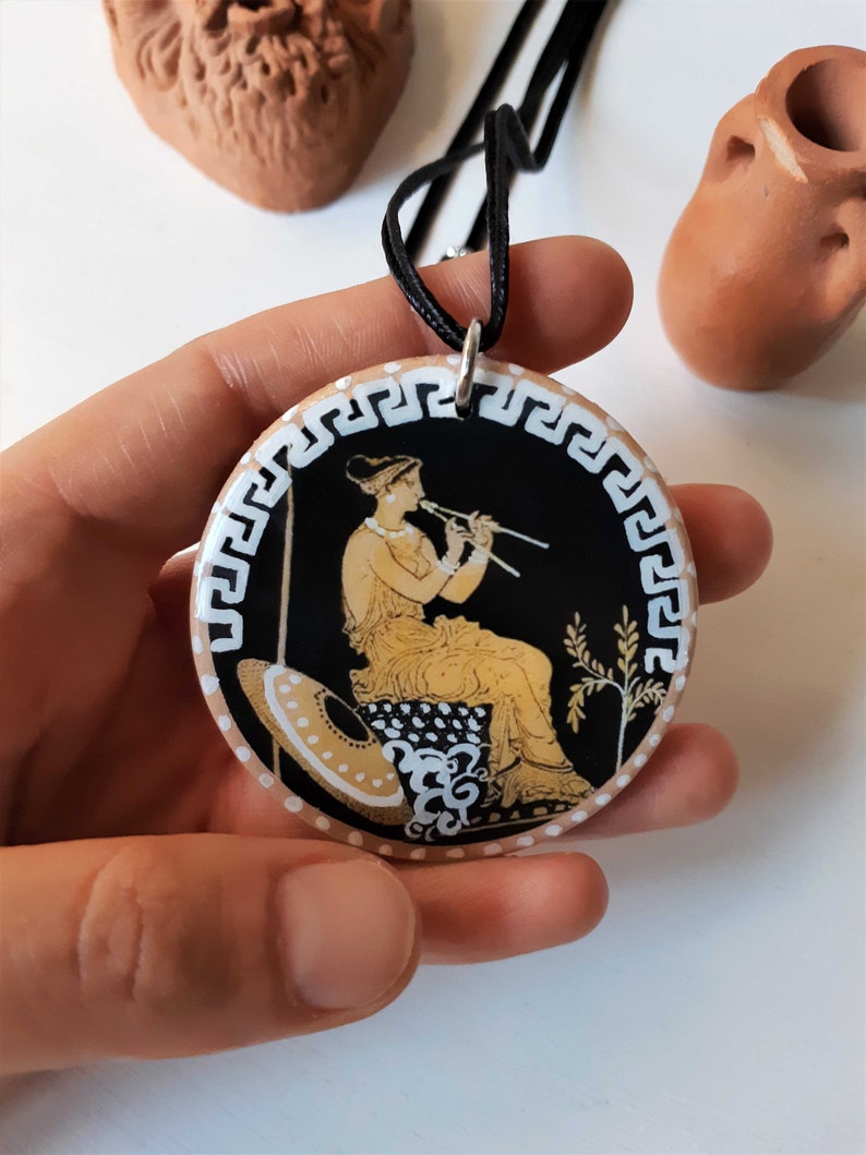 Peut inclure: Un collier pendentif rond avec un fond noir repr&eacute;sentant une sc&egrave;ne grecque classique d'une femme jouant de la fl&ucirc;te. Le pendentif a une bordure blanche avec un motif de cl&eacute; grecque et est sur un cordon noir.