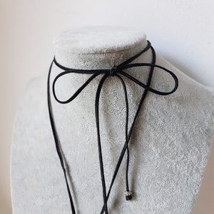 Peut inclure: Un collier ras du cou noir avec un d&eacute;tail en forme de n&oelig;ud. Le collier est pr&eacute;sent&eacute; sur un buste de mannequin gris. Le choker a un design d&eacute;licat et minimaliste, avec un cordon fin qui se noue dans le dos. Une petite perle argent&eacute;e pend au bout d'un des cordons.