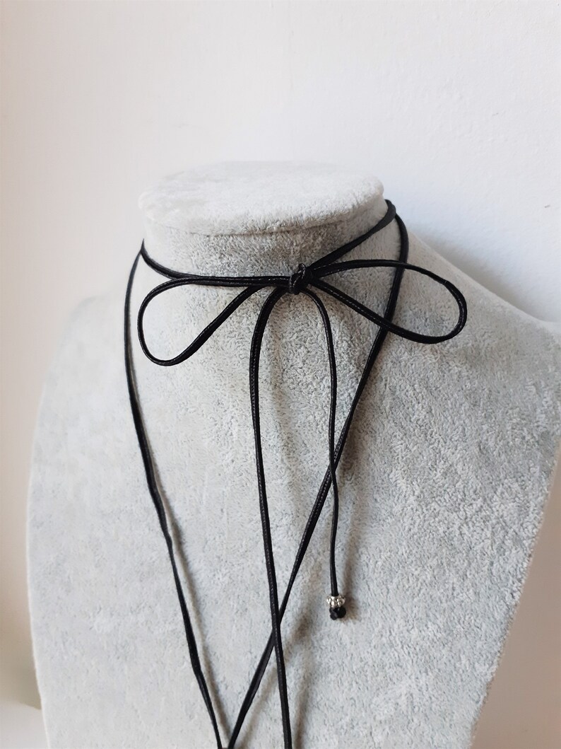 Peut inclure: Un collier ras du cou noir avec un d&eacute;tail en forme de n&oelig;ud. Le collier est pr&eacute;sent&eacute; sur un buste de mannequin gris. Le choker a un design d&eacute;licat et minimaliste, avec un cordon fin qui se noue dans le dos. Une petite perle argent&eacute;e pend au bout d'un des cordons.
