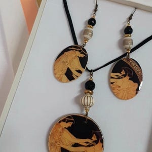 May include: A black necklace with a black cord and two oval pendants featuring black and gold Greek pottery designs. The necklace also has a round pendant with a similar design. The necklace comes with matching earrings with black and gold beads.