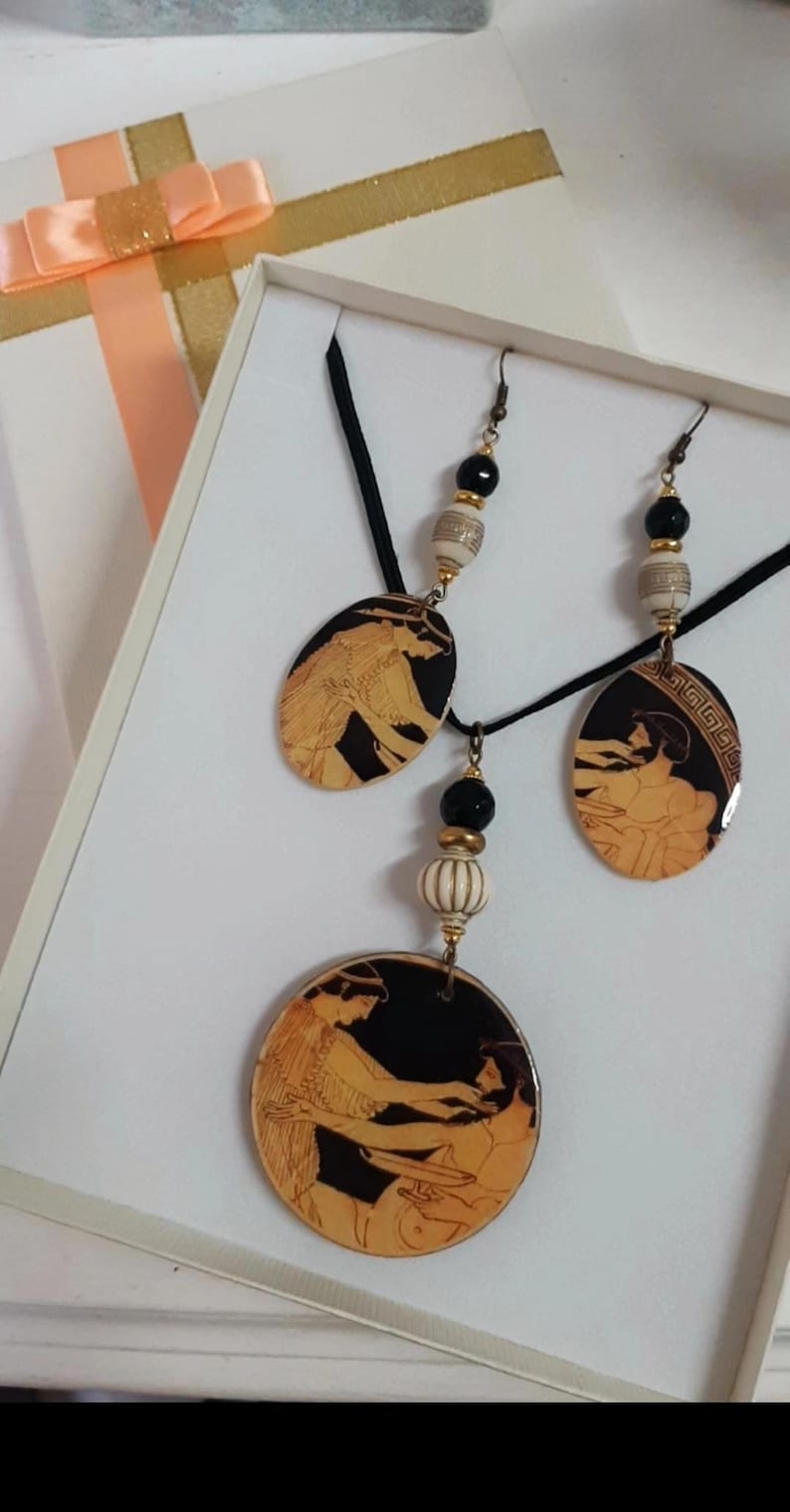 May include: A black necklace with a black cord and two oval pendants featuring black and gold Greek pottery designs. The necklace also has a round pendant with a similar design. The necklace comes with matching earrings with black and gold beads.