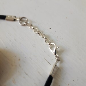 Puede incluir: Primer plano de un collar gargantilla negro con una cadena y un cierre plateados. El collar est&aacute; sobre una superficie blanca.