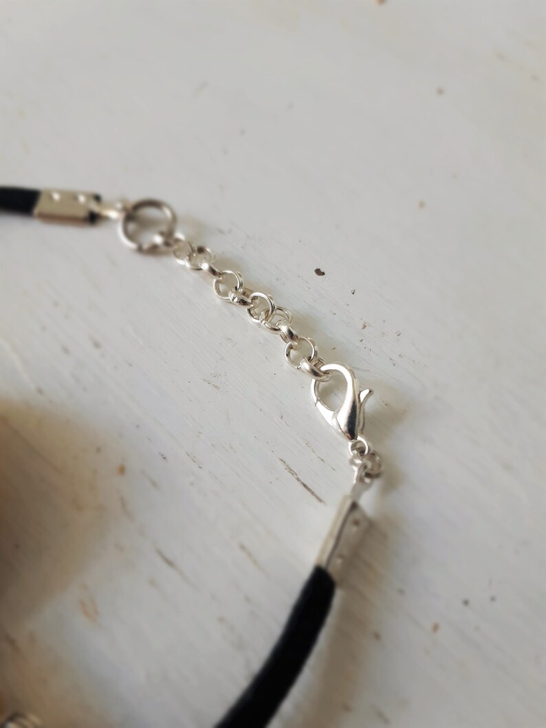 Puede incluir: Primer plano de un collar gargantilla negro con una cadena y un cierre plateados. El collar est&aacute; sobre una superficie blanca.