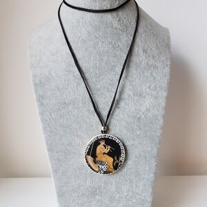 Peut inclure: Un collier &agrave; cordon noir avec un pendentif rond repr&eacute;sentant une sc&egrave;ne grecque classique. Le pendentif a un fond noir avec une figure jouant de la fl&ucirc;te, avec des d&eacute;tails dor&eacute;s et blancs.