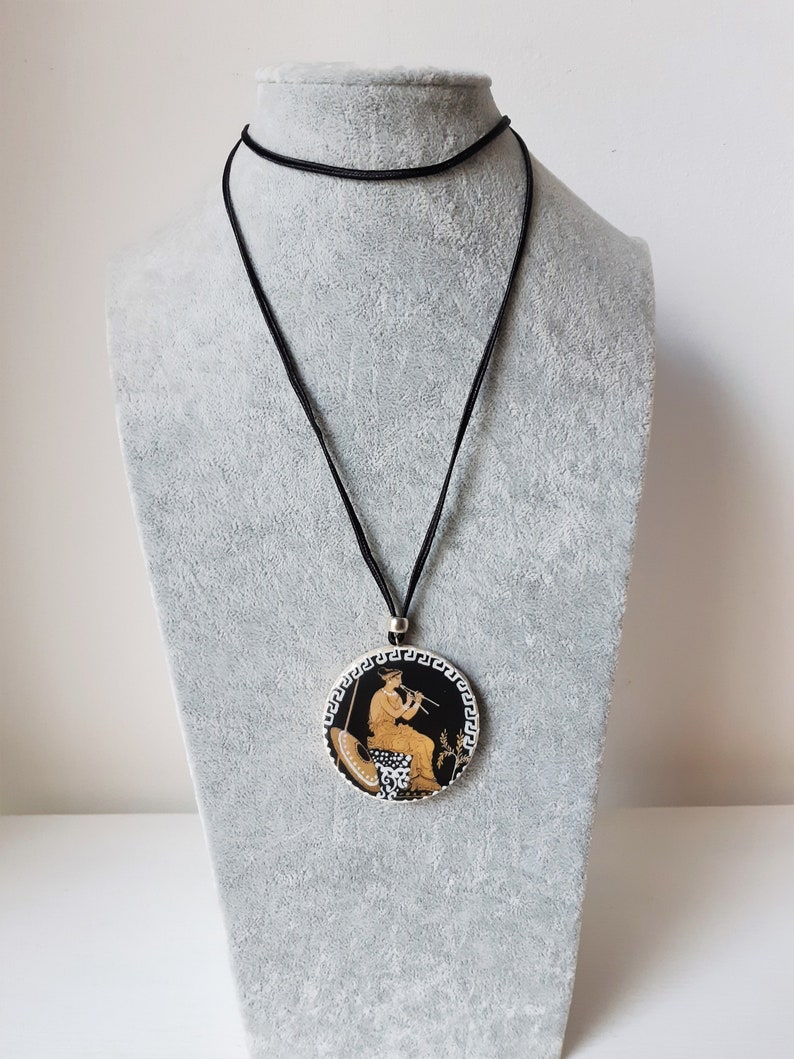 Peut inclure: Un collier &agrave; cordon noir avec un pendentif rond repr&eacute;sentant une sc&egrave;ne grecque classique. Le pendentif a un fond noir avec une figure jouant de la fl&ucirc;te, avec des d&eacute;tails dor&eacute;s et blancs.