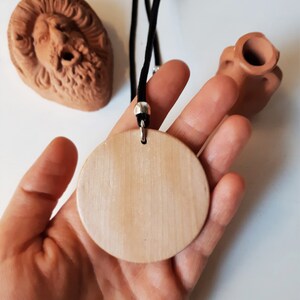 Peut inclure: Un collier avec un pendentif en bois clair et un cordon noir, tenu dans une main. Le pendentif est rond et lisse. En arri&egrave;re-plan, deux sculptures en terre cuite, un visage et un petit vase.