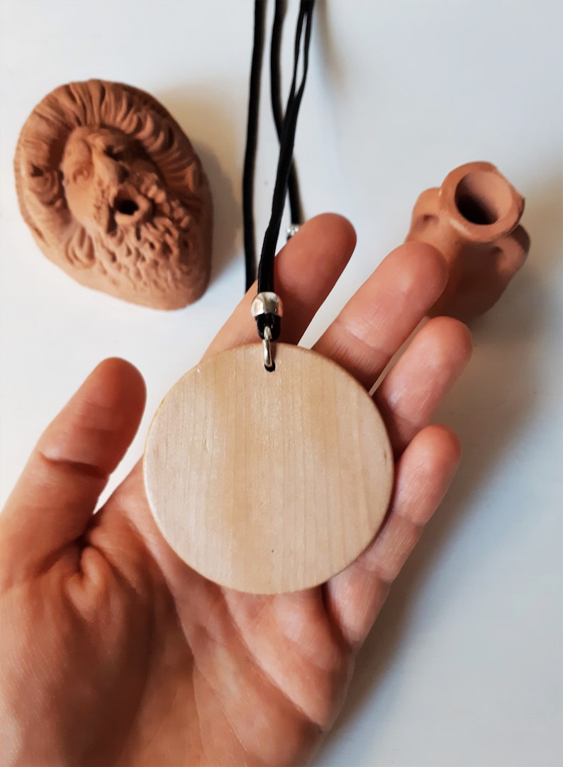 Peut inclure: Un collier avec un pendentif en bois clair et un cordon noir, tenu dans une main. Le pendentif est rond et lisse. En arri&egrave;re-plan, deux sculptures en terre cuite, un visage et un petit vase.
