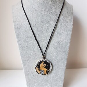 Peut inclure: Un collier pendentif rond avec un motif grec classique. Le pendentif pr&eacute;sente un fond noir avec une figure jouant de la fl&ucirc;te, entour&eacute;e d'une bordure blanche et noire. Le collier a un cordon noir.