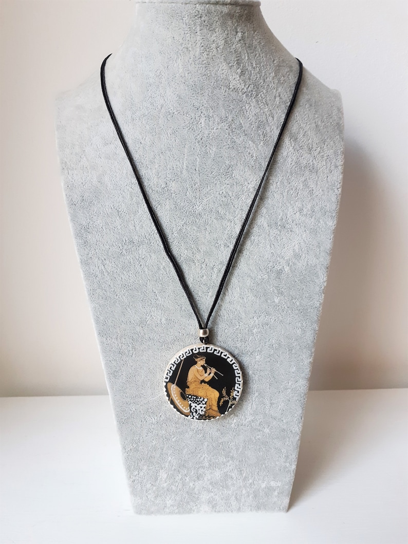 Peut inclure: Un collier pendentif rond avec un motif grec classique. Le pendentif pr&eacute;sente un fond noir avec une figure jouant de la fl&ucirc;te, entour&eacute;e d'une bordure blanche et noire. Le collier a un cordon noir.