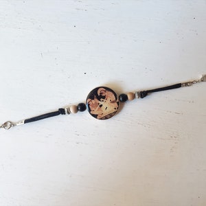 Puede incluir: Pulsera de cuero negro con un dije redondo de cer&aacute;mica que presenta una imagen en blanco y negro de una escena de la mitolog&iacute;a griega. La pulsera tiene cuentas de madera y un cierre plateado.