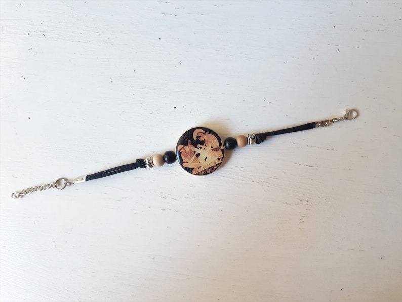 Puede incluir: Pulsera de cuero negro con un dije redondo de cer&aacute;mica que presenta una imagen en blanco y negro de una escena de la mitolog&iacute;a griega. La pulsera tiene cuentas de madera y un cierre plateado.