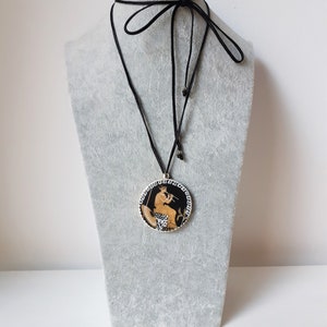 Peut inclure: Un collier &agrave; cordon noir avec un pendentif rond repr&eacute;sentant une sc&egrave;ne grecque classique. Le pendentif a un fond noir avec une figure jouant de la fl&ucirc;te, entour&eacute;e d'une bordure blanche et noire. Le collier est pr&eacute;sent&eacute; sur un mannequin gris.