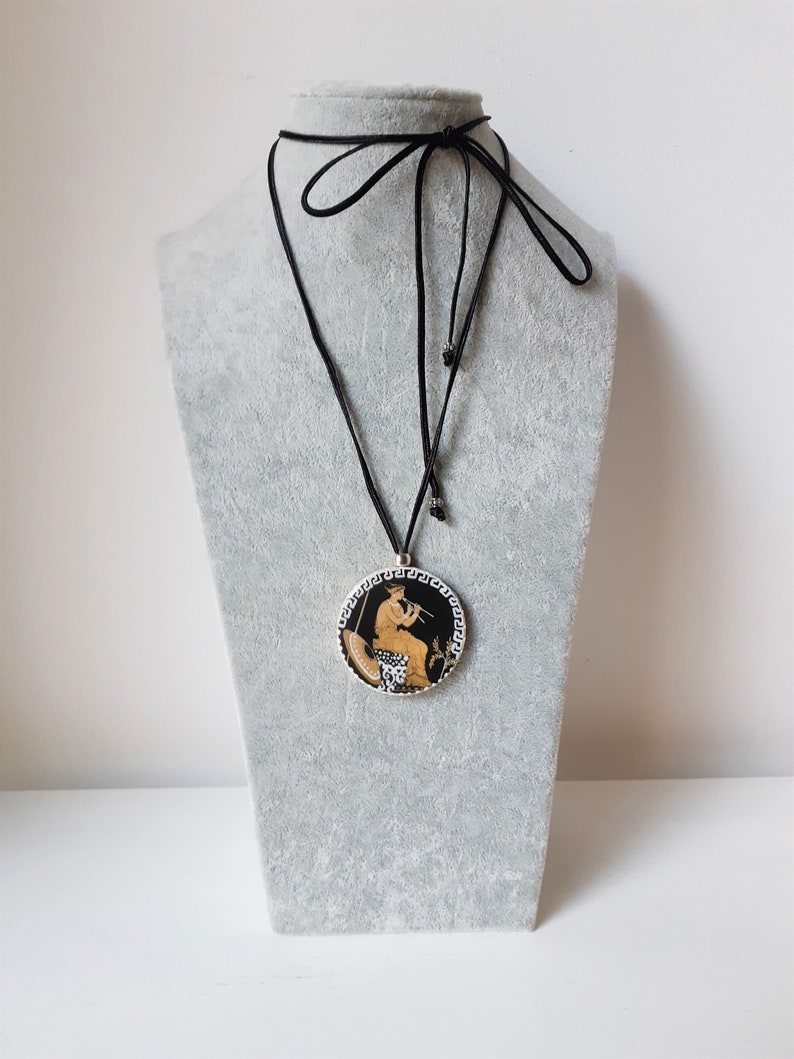 Peut inclure: Un collier &agrave; cordon noir avec un pendentif rond repr&eacute;sentant une sc&egrave;ne grecque classique. Le pendentif a un fond noir avec une figure jouant de la fl&ucirc;te, entour&eacute;e d'une bordure blanche et noire. Le collier est pr&eacute;sent&eacute; sur un mannequin gris.