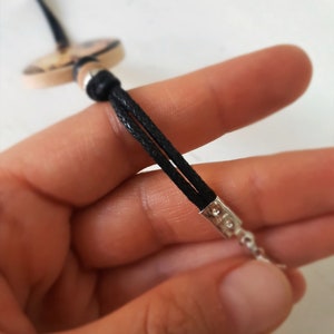 Puede incluir: Primer plano de un collar de cord&oacute;n negro con cierre plateado. El cord&oacute;n est&aacute; doblado y atado con una cuenta plateada. El collar est&aacute; sostenido en una mano.