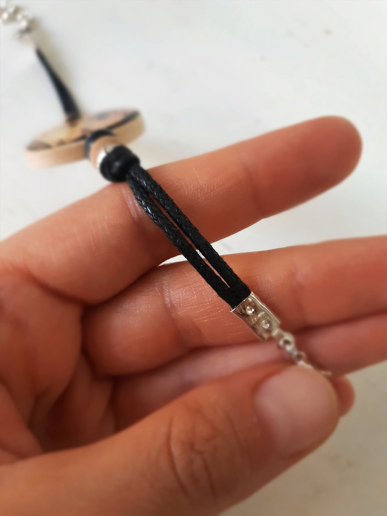 Puede incluir: Primer plano de un collar de cord&oacute;n negro con cierre plateado. El cord&oacute;n est&aacute; doblado y atado con una cuenta plateada. El collar est&aacute; sostenido en una mano.