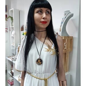 Peut inclure: Une femme porte une robe blanche avec des d&eacute;tails dor&eacute;s, une ceinture en corde dor&eacute;e et un ras du cou noir. Elle porte &eacute;galement un collier pendentif avec un motif circulaire et un bracelet manchette en cuivre. Un accessoire dor&eacute; en forme d'&eacute;pi de bl&eacute; est &eacute;pingl&eacute; &agrave; la robe.