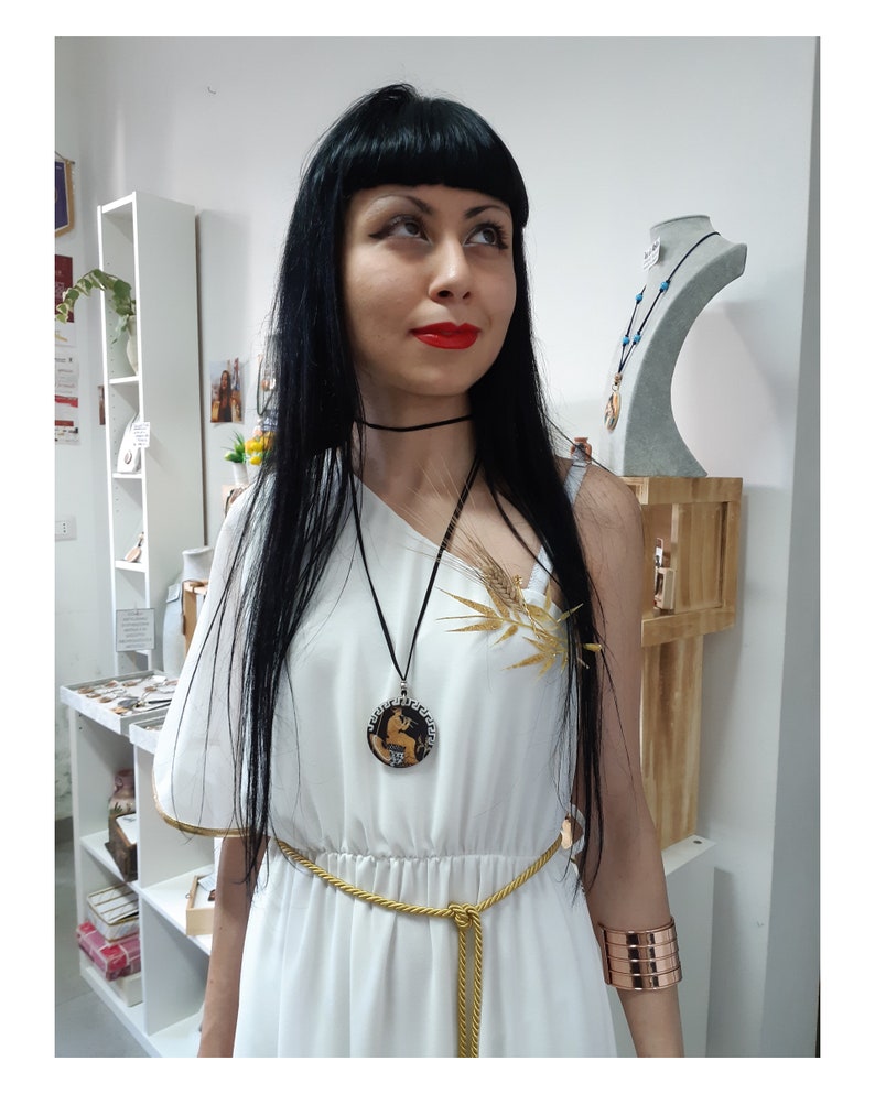 Peut inclure: Une femme porte une robe blanche avec des d&eacute;tails dor&eacute;s, une ceinture en corde dor&eacute;e et un ras du cou noir. Elle porte &eacute;galement un collier pendentif avec un motif circulaire et un bracelet manchette en cuivre. Un accessoire dor&eacute; en forme d'&eacute;pi de bl&eacute; est &eacute;pingl&eacute; &agrave; la robe.