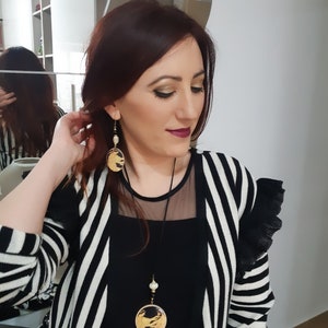 May include: A woman wearing a black and white striped cardigan with a black top and a gold necklace with a round pendant. She is also wearing gold earrings with round pendants.