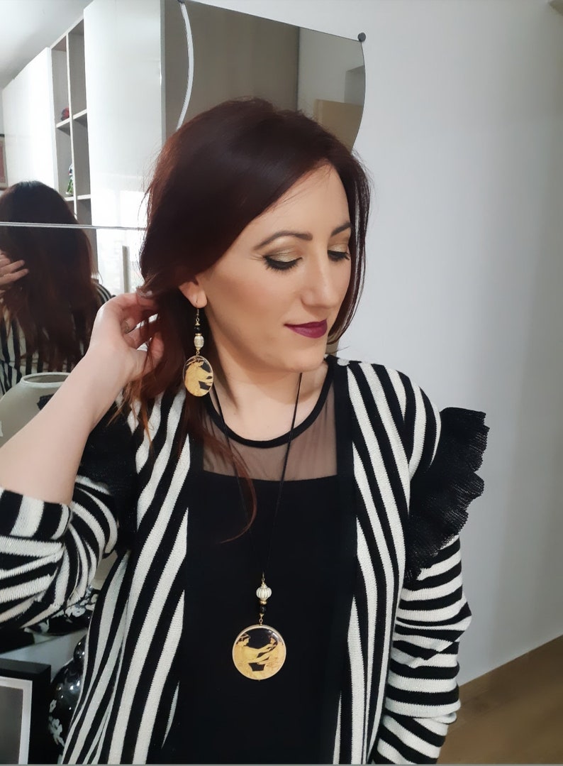 May include: A woman wearing a black and white striped cardigan with a black top and a gold necklace with a round pendant. She is also wearing gold earrings with round pendants.