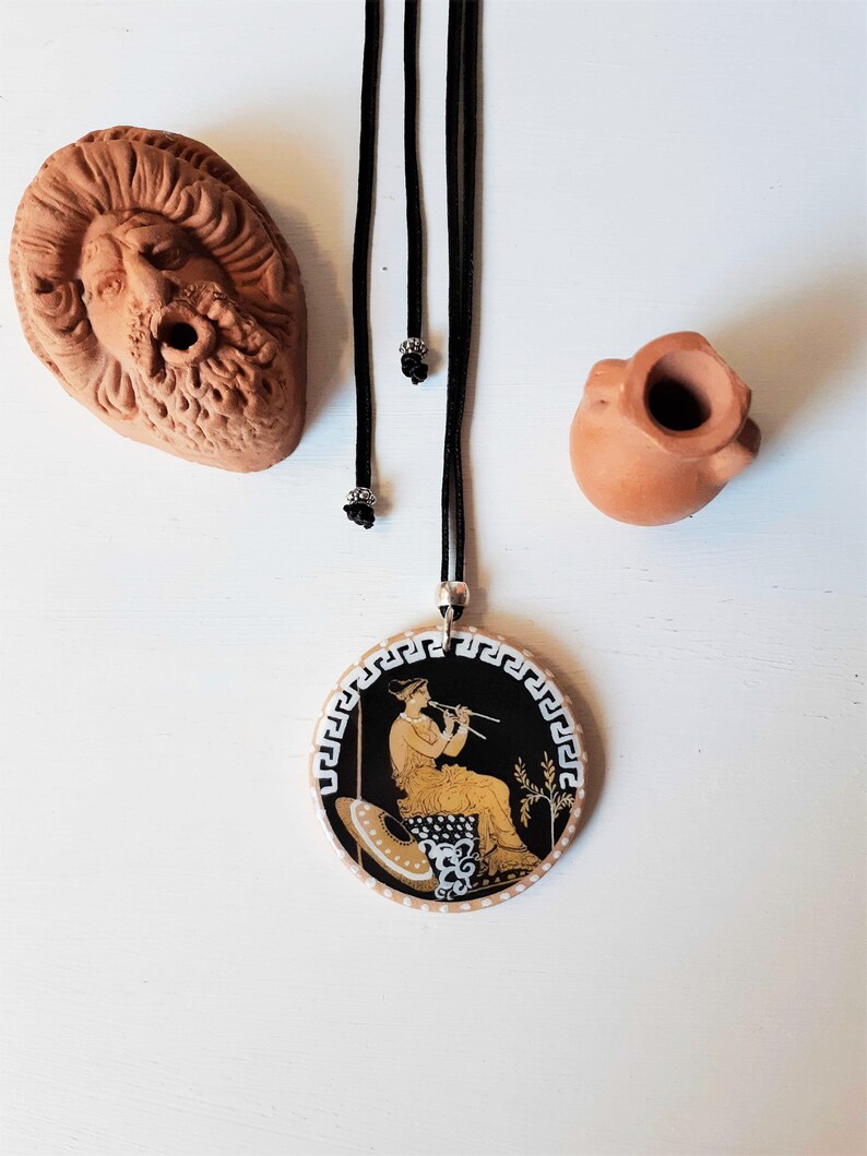 Peut inclure: Un collier avec un pendentif rond et un cordon noir. Le pendentif pr&eacute;sente une sc&egrave;ne grecque classique en or et noir. Deux petites sculptures en terre cuite et un visage en terre cuite sont &eacute;galement dans l'image.