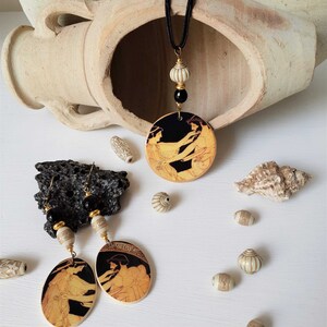May include: A black cord necklace with a black and gold pendant featuring a black and gold image of two women. The pendant is surrounded by white beads. The necklace is displayed on a white surface with white beads and a seashell.