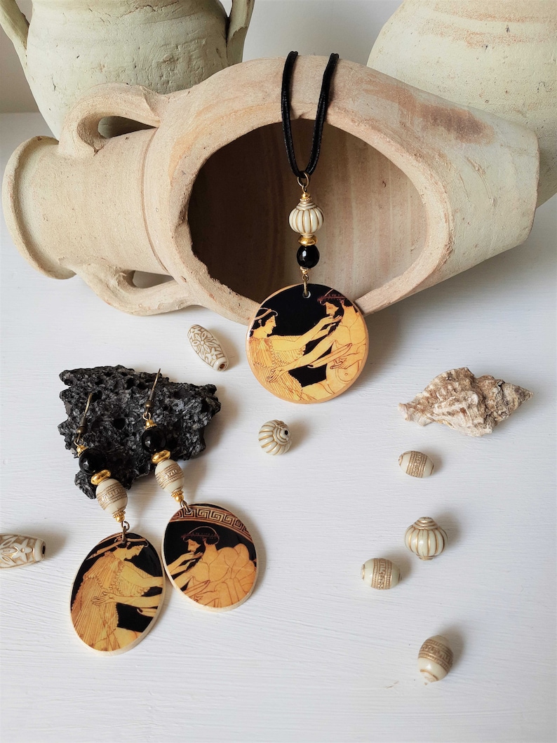 May include: A black cord necklace with a black and gold pendant featuring a black and gold image of two women. The pendant is surrounded by white beads. The necklace is displayed on a white surface with white beads and a seashell.