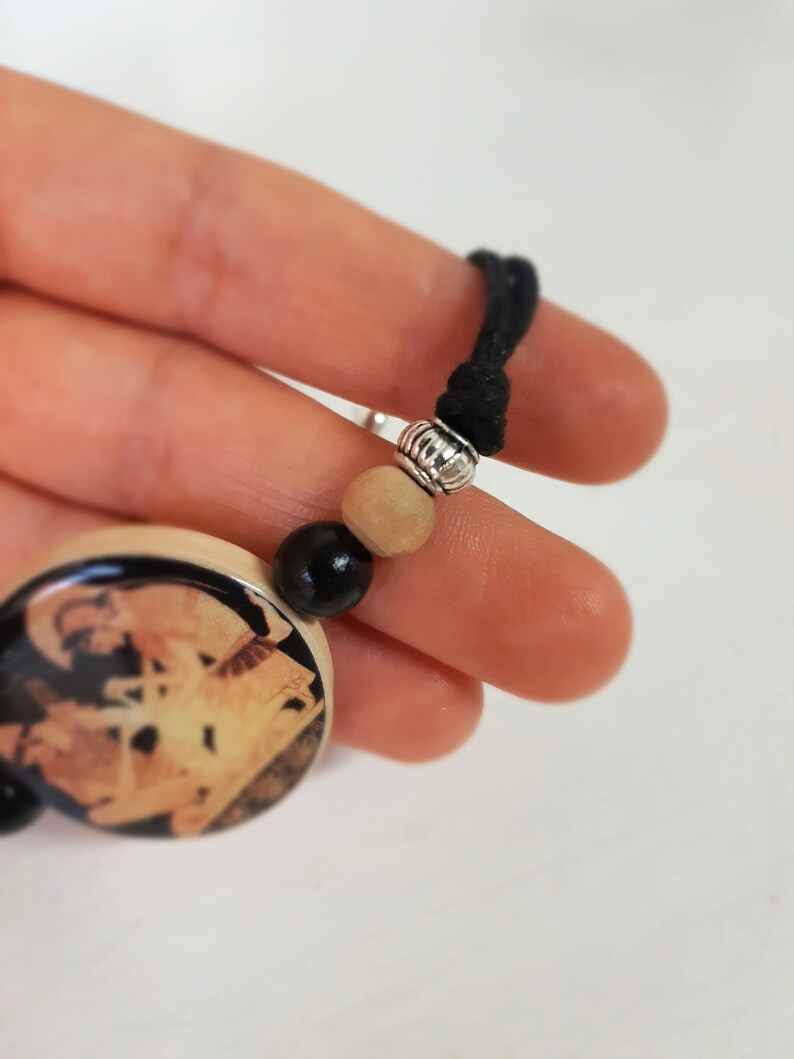 Puede incluir: Un amuleto de madera con una imagen en blanco y negro de un edificio, unido a un cord&oacute;n negro con cuentas de madera y plata.