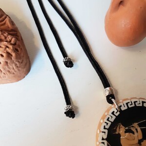 Peut inclure: Un collier &agrave; cordon noir avec un pendentif repr&eacute;sentant une illustration noire et beige d'une personne jouant de la fl&ucirc;te. Le pendentif a une bordure blanche avec un motif de cl&eacute; grecque. Le collier a des accents m&eacute;talliques argent&eacute;s.
