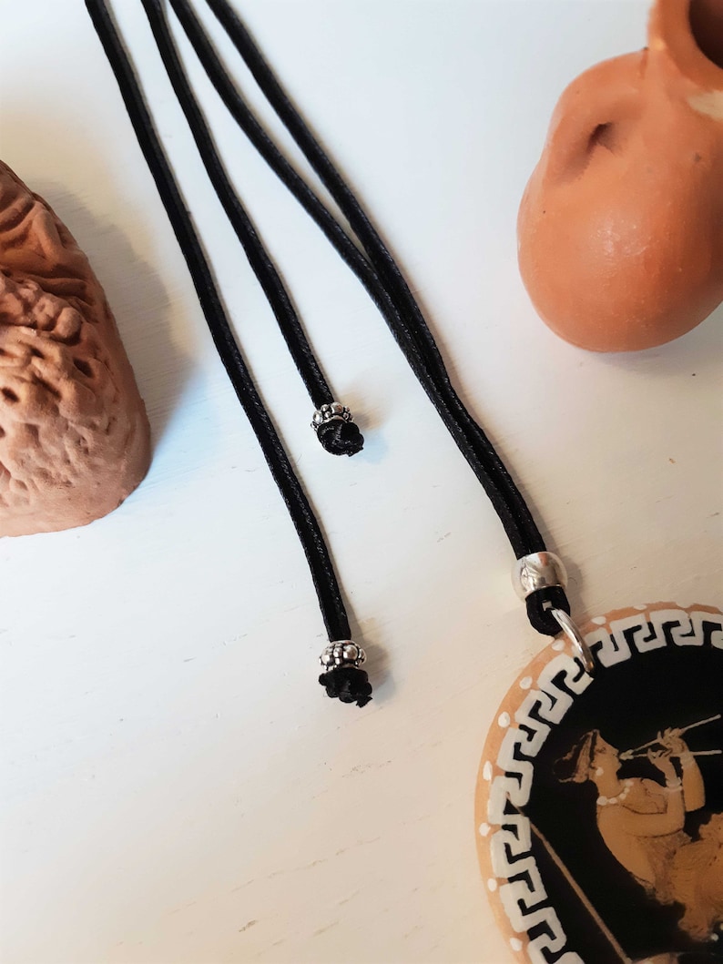 Peut inclure: Un collier &agrave; cordon noir avec un pendentif repr&eacute;sentant une illustration noire et beige d'une personne jouant de la fl&ucirc;te. Le pendentif a une bordure blanche avec un motif de cl&eacute; grecque. Le collier a des accents m&eacute;talliques argent&eacute;s.