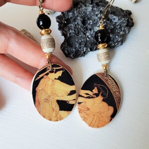 May include: A pair of dangle earrings with black and white ceramic beads and oval-shaped pendants featuring a black and gold Greek vase design.