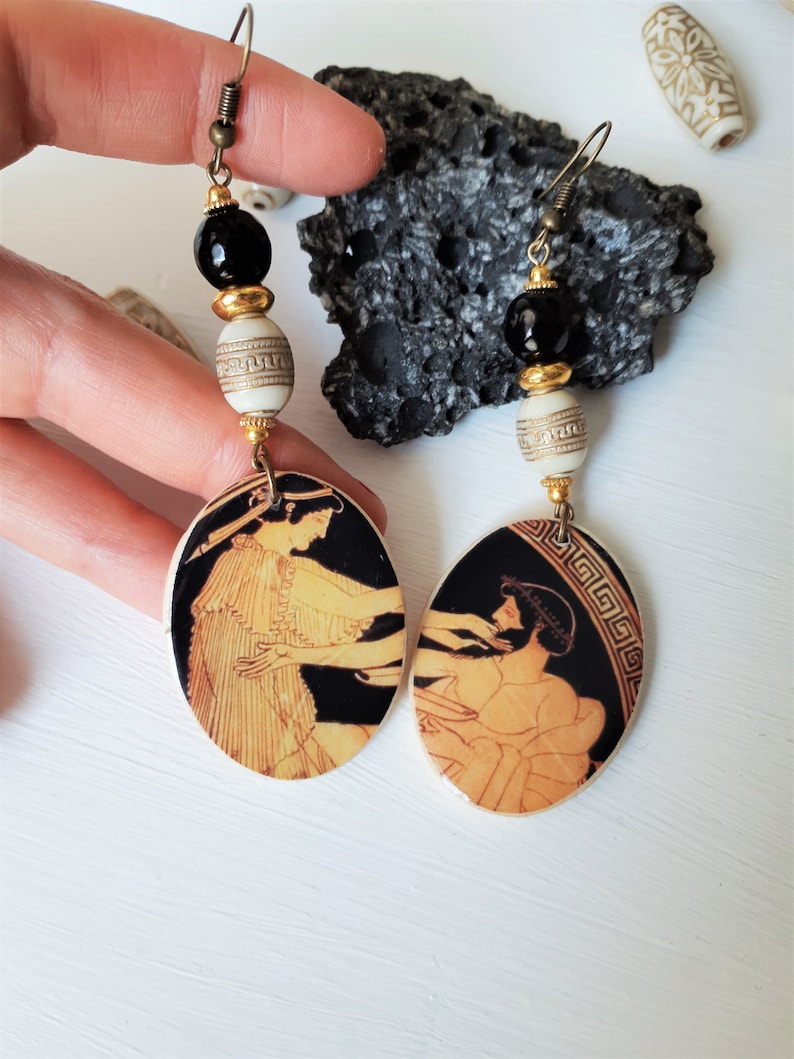 May include: A pair of dangle earrings with black and white ceramic beads and oval-shaped pendants featuring a black and gold Greek vase design.