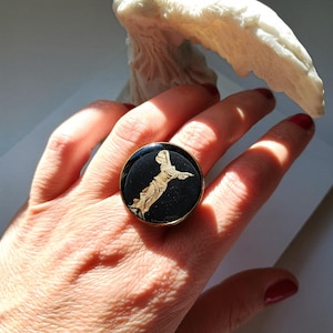 May include: A gold-toned ring with a black face featuring a gold-colored statue design. The ring is round and has a detailed image of a classical figure. The ring is on a person's finger.